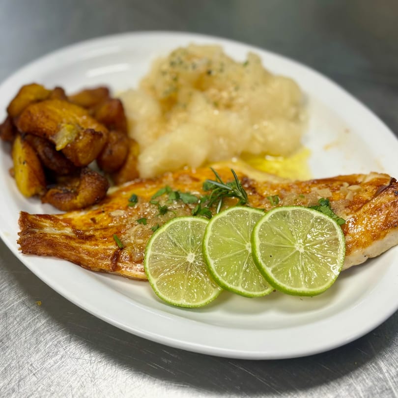 Cuban-style grilled fish with lime slices, plantains, and mashed yuca on a white plate.