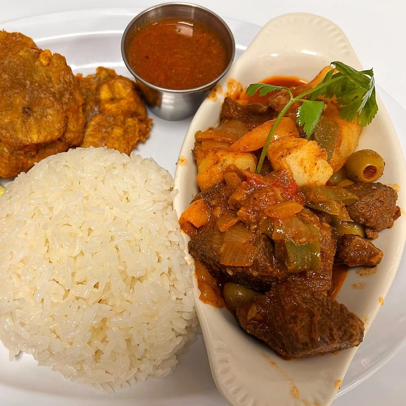 Cuban Ropa Vieja with white rice, fried plantains, and a side of red sauce on a white plate.
